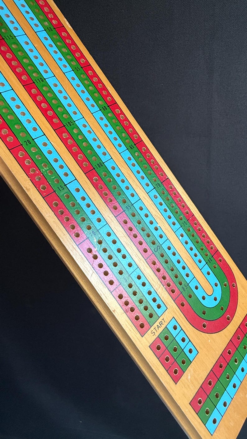 May include: Wooden cribbage board with red, green, and blue scoring tracks. The board has a rectangular shape with a curved end and a small square area labeled "START". The board is made of light-colored wood.
