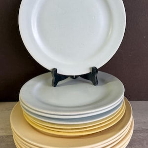 May include: A stack of ceramic plates in various colors, including white, light blue, and yellow. The plates are displayed on a wooden surface, with one white plate elevated on a black stand. The plates are round with a slightly raised edge.