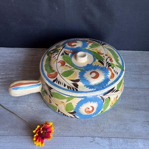 May include: A ceramic soup pot with a lid, featuring a hand-painted floral design in blue, green, and orange on a cream background. The pot has a handle and a small knob on the lid. The pot is on a wooden surface.