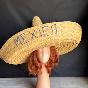 May include: A large straw sombrero, woven with a wide brim and a tall crown. The word "MEXICO" is embroidered in blue thread across the brim. The hat is displayed on a mannequin head with red hair.