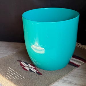 May include: A turquoise-colored, cylindrical planter with a rounded base. The planter sits on a small, rectangular, patterned rug with a Native American design. The rug has a gray background with red, white, and black accents.
