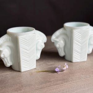 May include: Two white, art deco-style candle holders. Each holder features a central rectangular column with a chevron design and two horse heads on either side. A small sprig of purple flowers rests in front of the candle holders.