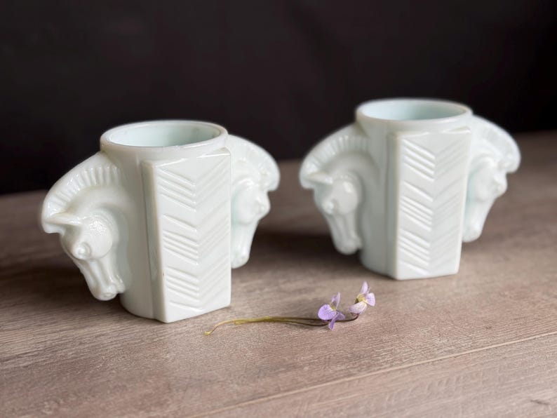 May include: Two white, art deco-style candle holders. Each holder features a central rectangular column with a chevron design and two horse heads on either side. A small sprig of purple flowers rests in front of the candle holders.