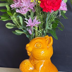 May include: A glossy orange ceramic squirrel figurine holding an acorn, serving as a vase filled with a vibrant bouquet of yellow sunflowers, red carnations, pink chrysanthemums, and small yellow and white filler flowers against a black background.