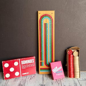 May include: A collection of classic board games and card games, including a red dominoes box, a wooden cribbage board, a pink card box, and a box of red and tan game pieces. The dominoes box reads "DOMINOES".