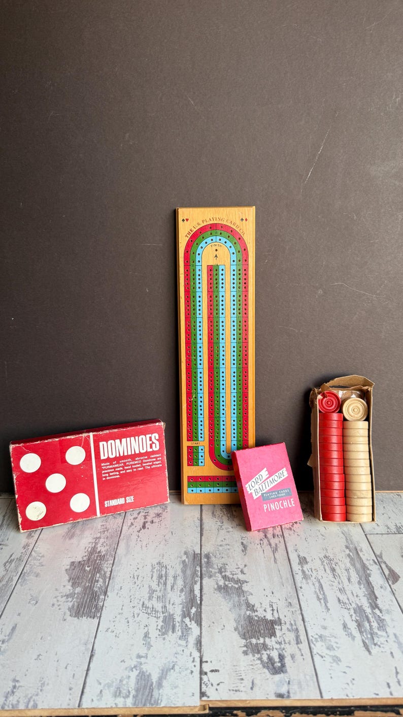 May include: A collection of classic board games and card games, including a red dominoes box, a wooden cribbage board, a pink card box, and a box of red and tan game pieces. The dominoes box reads "DOMINOES".