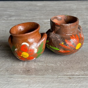 May include: Two small, hand-painted terracotta pots. The pots are brown with colorful floral designs in red, orange, yellow, and green. The pots have a rustic, handmade appearance, with a slightly rough texture.