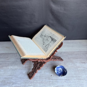 May include: An open antique book with illustrations, resting on a carved wooden book stand. The stand is a dark reddish-brown color. A small, blue and white decorative bowl sits nearby. The book's pages are aged and show signs of wear.
