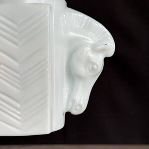 May include: A white, art deco style ceramic mug with a horse head design on the handle. The mug features a geometric pattern on the side and is set against a dark background.