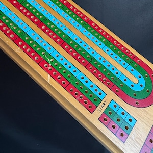 May include: Wooden cribbage board with red, green, and blue tracks. The board has a curved design with a start area and pegging holes. The word "START" is printed on the board.