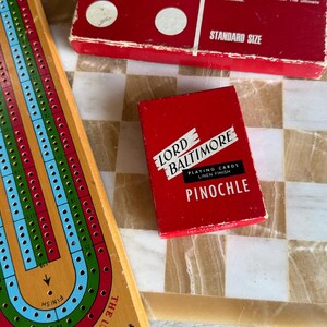 May include: A wooden cribbage board, a red box of dominoes, and a red box of Lord Baltimore Pinochle playing cards are arranged on a checkered surface. The dominoes box has white dots and the text "DOMINOES". The playing cards box has the text "PINOCHLE".