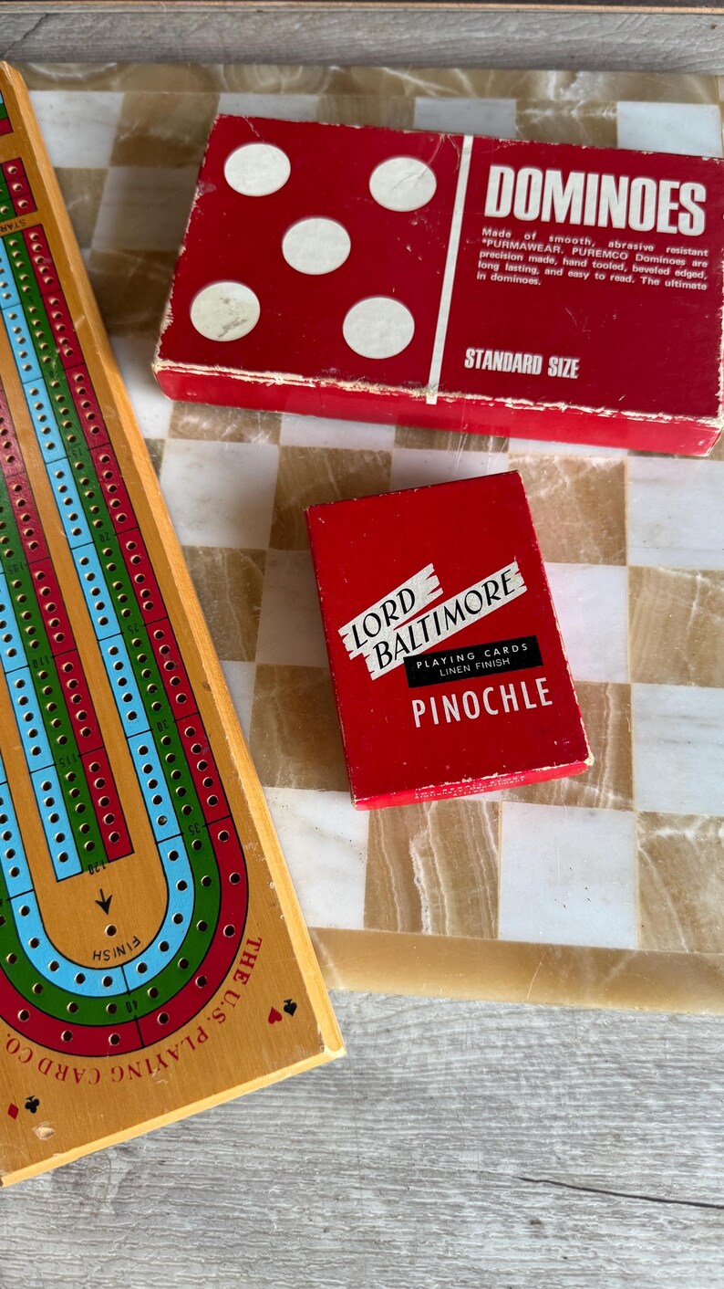 May include: A wooden cribbage board, a red box of dominoes, and a red box of Lord Baltimore Pinochle playing cards are arranged on a checkered surface. The dominoes box has white dots and the text "DOMINOES". The playing cards box has the text "PINOCHLE".