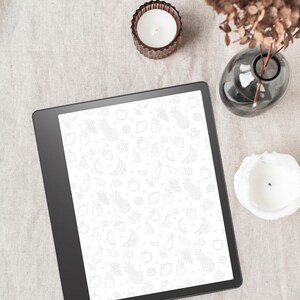May include: A black and white digital tablet with a white background and a pattern of fruits and vegetables. The tablet is on a light brown surface with a white candle and a gray vase.