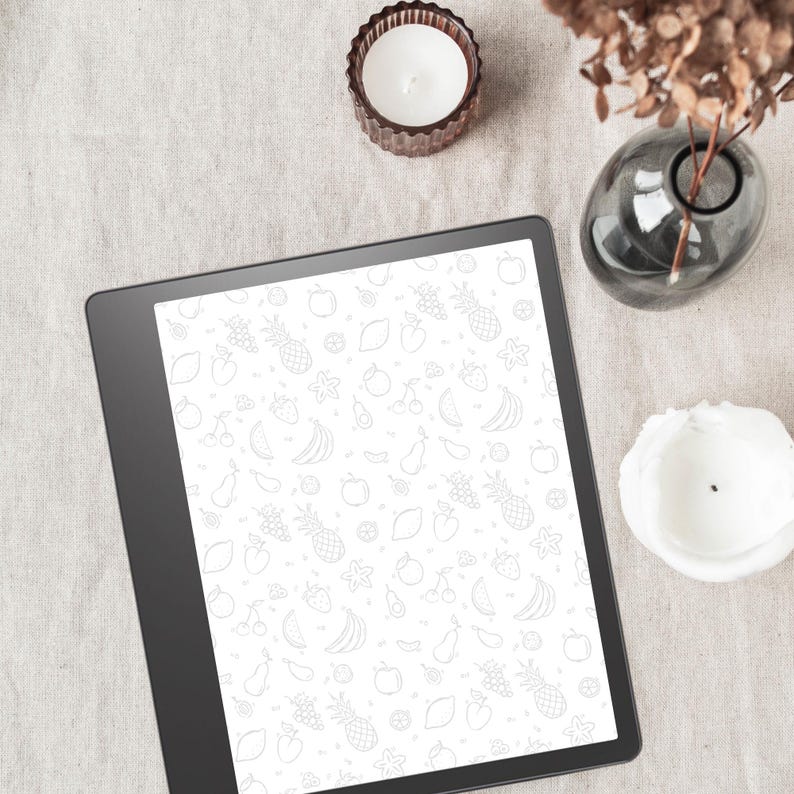 May include: A black and white digital tablet with a white background and a pattern of fruits and vegetables. The tablet is on a light brown surface with a white candle and a gray vase.