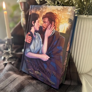 May include: A hardcover book titled Outlander by Diana Gabaldon featuring an illustrated cover of a couple embracing. The artwork displays a woman and a man with reddish-brown hair in a warm, sunset-lit setting. The title is printed in gold lettering.