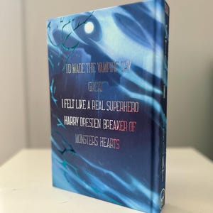 Pode incluir: Um livro de capa dura com uma capa azul com o texto "ID MADE THE VAMPIAS CHY GREAT I FELT LIKE A REAL SUPERHERO HARRY DRESDEN BREAKER OF MONSTERS HEARTS." O livro tem um design de folha de prata e um fundo abstrato azul e branco.