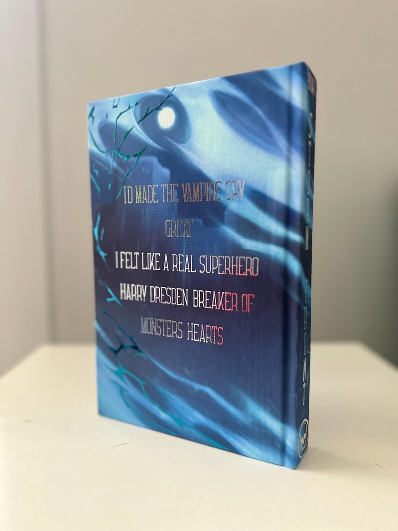 Pode incluir: Um livro de capa dura com uma capa azul com o texto "ID MADE THE VAMPIAS CHY GREAT I FELT LIKE A REAL SUPERHERO HARRY DRESDEN BREAKER OF MONSTERS HEARTS." O livro tem um design de folha de prata e um fundo abstrato azul e branco.