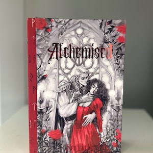 以下が含まれることがあります： ロマンチックなイラストが描かれた「Alchemist I」というタイトルのハードカバー本。表紙には、男性と女性、赤いバラ、赤い箔でタイトルが描かれたグレースケールのゴシック様式のシーンが描かれています。背表紙は赤色で、金色のアクセントが付いています。