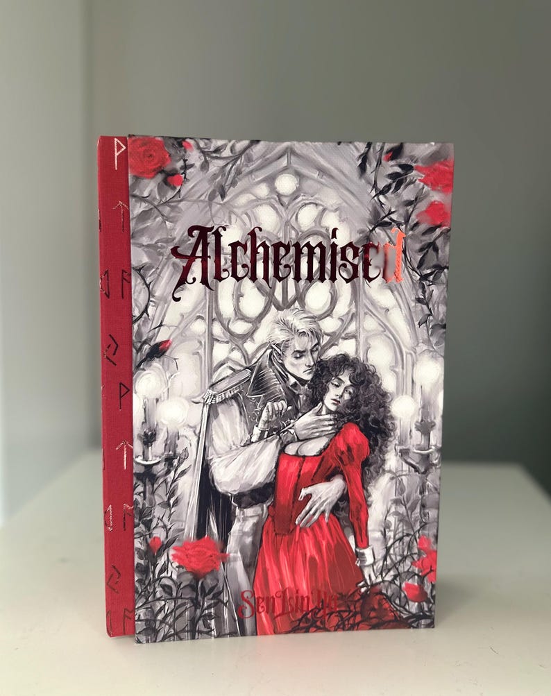 以下が含まれることがあります： ロマンチックなイラストが描かれた「Alchemist I」というタイトルのハードカバー本。表紙には、男性と女性、赤いバラ、赤い箔でタイトルが描かれたグレースケールのゴシック様式のシーンが描かれています。背表紙は赤色で、金色のアクセントが付いています。