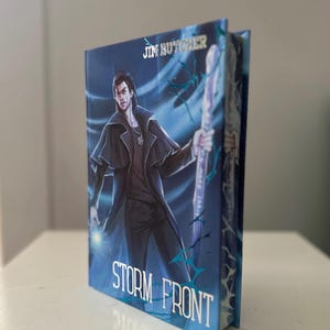 May include: A hardcover book titled "Storm Front" by Jim Butcher. The cover features a male character in a dark coat, holding a staff, with a blue and white background. The book is standing upright.