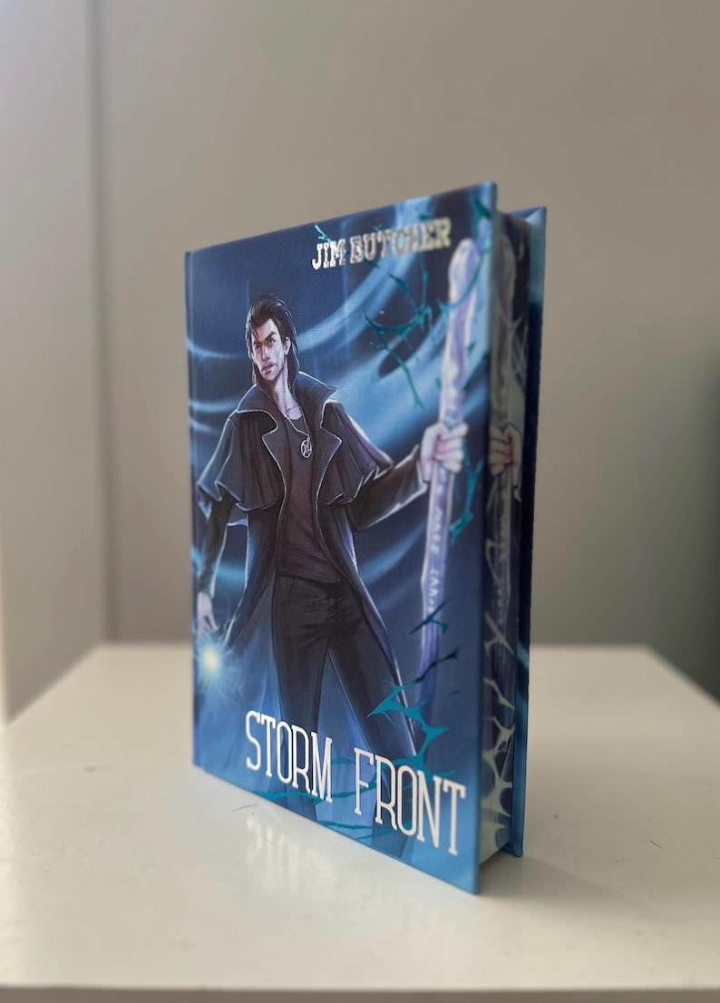Pode incluir: Um livro de capa dura intitulado "Storm Front" de Jim Butcher. A capa apresenta um personagem masculino com um casaco escuro, segurando uma bengala, sobre um fundo azul e branco. O livro est&aacute; em p&eacute;.