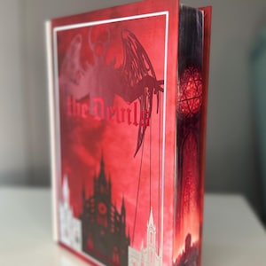 May include: A red book titled "the Devil" in gothic script. The cover displays a devil silhouette with wings and a cityscape. The spine features a stained glass window design.