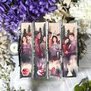 Könnte beinhalten: Vier nebeneinander angeordnete Bücher, die auf den Buchschnitten eine durchgehende Illustration zeigen. Das Bild zeigt menschliche Figuren in einer Waldlandschaft sowie Symbole wie einen roten Apfel, eine Blume, ein Band und eine weiße Schachfigur.