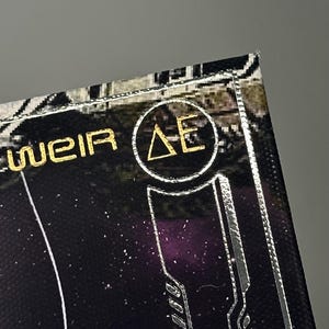 Puede incluir: Primer plano de un dise&ntilde;o negro y dorado con el texto "WEIR" y un logotipo circular con las letras "&Delta;E". El dise&ntilde;o tiene una est&eacute;tica futurista con acentos plateados y un fondo estrellado.