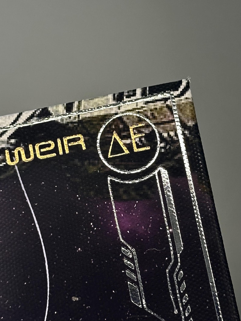 Puede incluir: Primer plano de un dise&ntilde;o negro y dorado con el texto "WEIR" y un logotipo circular con las letras "&Delta;E". El dise&ntilde;o tiene una est&eacute;tica futurista con acentos plateados y un fondo estrellado.