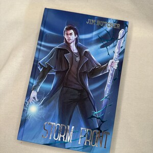 Pode incluir: Um livro de capa dura intitulado "Storm Front" de Jim Butcher. A capa apresenta uma ilustra&ccedil;&atilde;o de um homem com um casaco preto segurando uma bengala e um orbe brilhante, em um fundo azul com texto prateado.