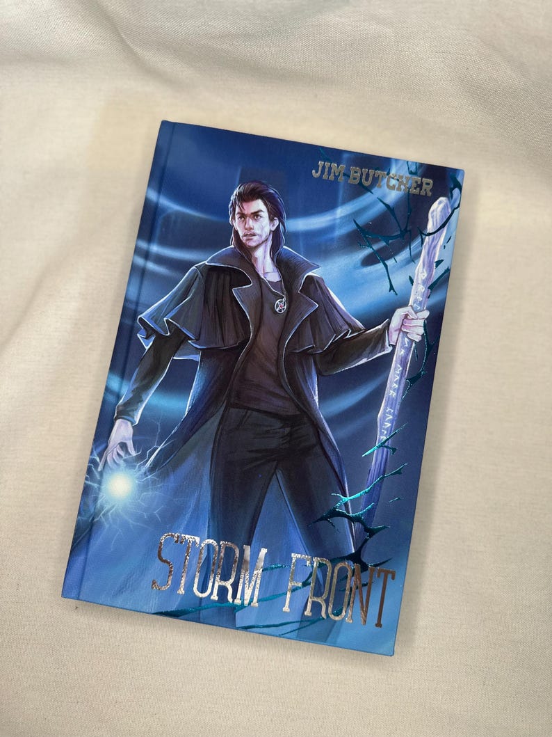 Pode incluir: Um livro de capa dura intitulado "Storm Front" de Jim Butcher. A capa apresenta uma ilustra&ccedil;&atilde;o de um homem com um casaco preto segurando uma bengala e um orbe brilhante, em um fundo azul com texto prateado.
