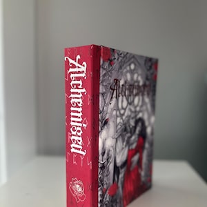 以下が含まれることがあります： 「Alchemised」というタイトルの本。赤い背表紙と表紙。背表紙には白いタイトル、バラ、著者の名前が記載されています。表紙は、赤いドレスを着た女性のグレースケールイラストです。