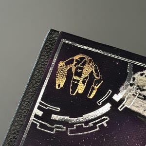 Puede incluir: Una secci&oacute;n de una cubierta de libro p&uacute;rpura oscuro con un dise&ntilde;o de mano dorada y detalles plateados. La cubierta tiene un borde negro similar al cuero. El fondo es un dise&ntilde;o espacial estrellado.