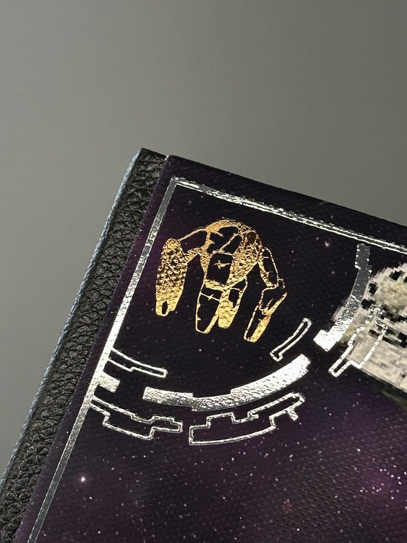 Puede incluir: Una secci&oacute;n de una cubierta de libro p&uacute;rpura oscuro con un dise&ntilde;o de mano dorada y detalles plateados. La cubierta tiene un borde negro similar al cuero. El fondo es un dise&ntilde;o espacial estrellado.