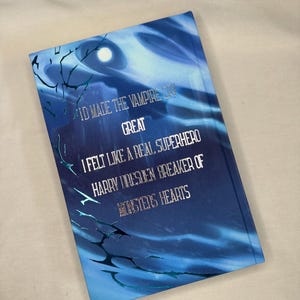 Pode incluir: Um livro de capa dura azul com o texto "I'D MADE THE VAMPIRE CRY GREAT I FELT LIKE A REAL SUPERHERO HARRY DRESDEN BREAKER OF MONSTERS HEARTS". A capa apresenta um design de c&eacute;u noturno com uma lua cheia e galhos de &aacute;rvores.