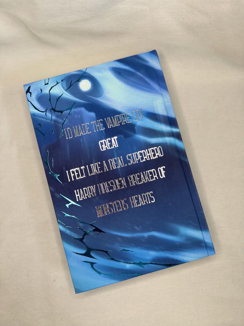 Pode incluir: Um livro de capa dura azul com o texto "I'D MADE THE VAMPIRE CRY GREAT I FELT LIKE A REAL SUPERHERO HARRY DRESDEN BREAKER OF MONSTERS HEARTS". A capa apresenta um design de c&eacute;u noturno com uma lua cheia e galhos de &aacute;rvores.