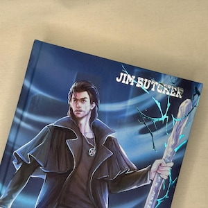 Pode incluir: Um livro de capa dura com uma capa azul com uma ilustra&ccedil;&atilde;o de um homem de casaco preto segurando uma bengala. O nome do autor, "JIM BUTCHER", est&aacute; impresso em prata. A obra de arte tem um tema de fantasia.
