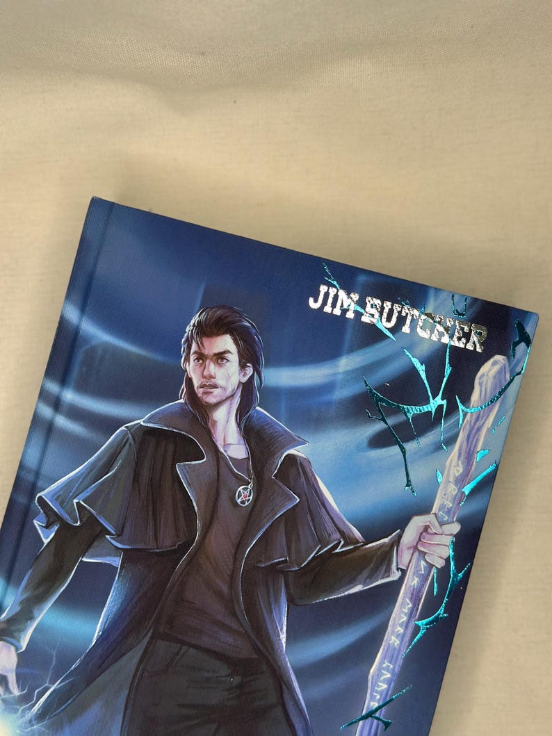 Pode incluir: Um livro de capa dura com uma capa azul com uma ilustra&ccedil;&atilde;o de um homem de casaco preto segurando uma bengala. O nome do autor, "JIM BUTCHER", est&aacute; impresso em prata. A obra de arte tem um tema de fantasia.