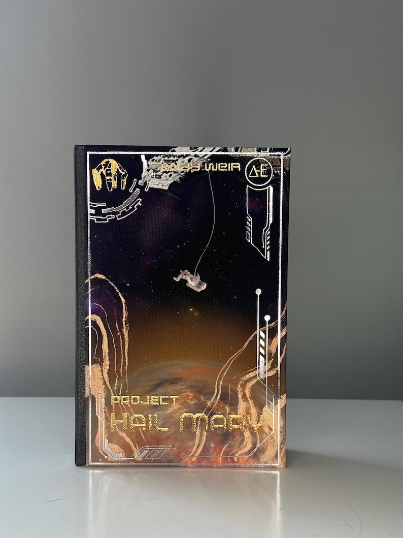 May include: Hardback book titled "Project Hail Mary" with a space theme. The cover shows an astronaut, a planet, and gold and white geometric designs. The author's name, Andy Weir, is also visible.