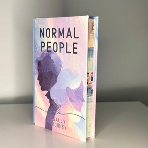 Op de afbeelding: Een hardcover boek getiteld "NORMAL PEOPLE" van Sally Rooney. De omslag toont een pastelkleurige illustratie van twee silhouetten, een man en een vrouw, met een hartsymbool. De rug van het boek toont een kleurrijke afbeelding van mensen.