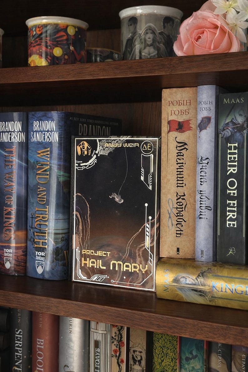 Puede incluir: Una estanter&iacute;a de madera llena de libros de tapa dura. Los t&iacute;tulos incluyen "Project Hail Mary", "Wind and Truth" y "The Way of Kings". Una taza decorativa es visible en el estante superior.