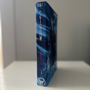 Pode incluir: Um livro de capa dura intitulado "Storm Front, Book 1" com uma capa azul. A lombada apresenta o t&iacute;tulo em texto vertical branco. Um &iacute;cone de caveira est&aacute; na parte inferior. O livro est&aacute; sobre uma superf&iacute;cie branca.