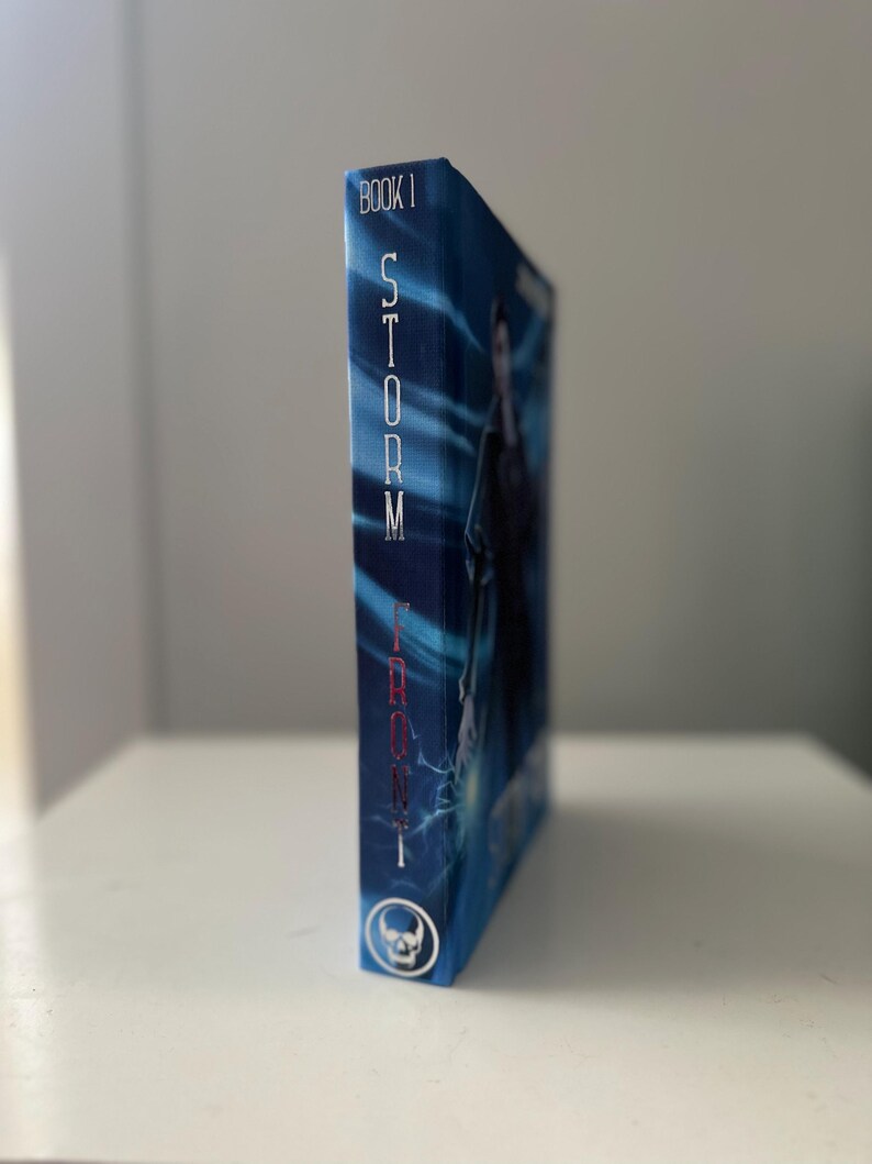 Pode incluir: Um livro de capa dura intitulado "Storm Front, Book 1" com uma capa azul. A lombada apresenta o t&iacute;tulo em texto vertical branco. Um &iacute;cone de caveira est&aacute; na parte inferior. O livro est&aacute; sobre uma superf&iacute;cie branca.