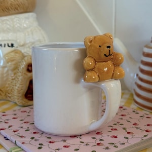 Puede incluir: Una taza de cerámica blanca con una figura de oso de peluche marrón adherida al borde. La taza está sobre un cuaderno rosa con estampado floral. Otros artículos de cerámica son visibles en el fondo.