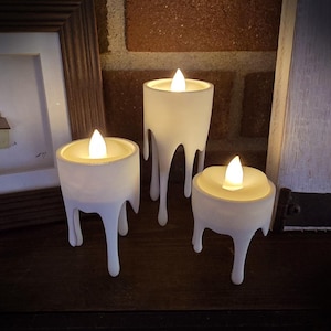 May include: Three white, drippy candle holders with LED lights. The candle holders are different sizes and have a melted wax appearance. The background includes a brick wall and a framed picture.