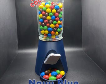 May include: A navy blue candy dispenser with a clear glass top filled with multi-colored chocolate candies. The dispenser features a dispensing lever and a small tray at the base containing loose candies. Text reads NEW COLOR and Navy Blue.