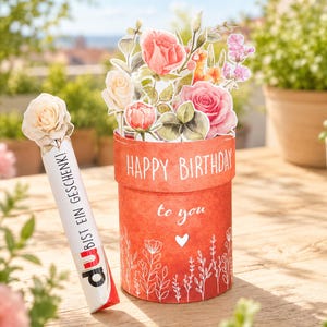 May include: A cylindrical red gift box featuring the white text HAPPY BIRTHDAY to you and a small heart icon, topped with a paper floral arrangement of pink and cream roses. Beside it stands a white tube with the text DU BIST EIN GESCHENK!