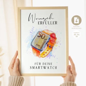 May include: A framed print with a watercolour illustration of a smartwatch, displaying a 50 Euro note on its screen. The watch has an orange strap. The text on the print reads "Wunsch Erfüller" and "Für deine Smartwatch".