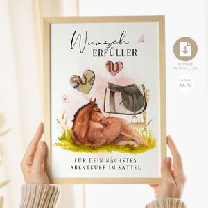 Op de afbeelding: Een ingelijste aquarelprint met een bruin paard dat in het gras ligt, een zadel en de tekst "Wunsch Erfüller" en "Für dein nächstes Abenteuer im Sattel." Twee hartvormige ballonnen tonen de nummers 50 en 10. De print wordt door handen vastgehouden.