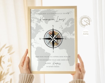 May include: A framed print with a light wood frame. The print features a world map background with a compass rose design. Text includes "Kommunion, Louis!" and inspirational phrases. The print is held by a person.